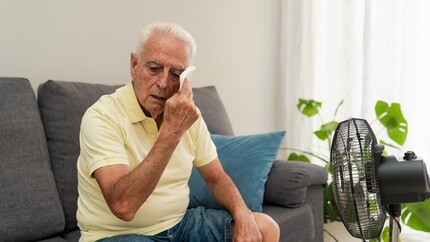 oudere man dept zijn gezicht zittend op de bank met ventilator op zich gericht